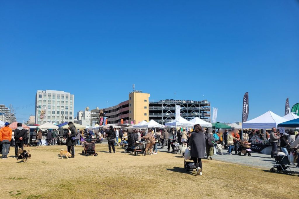 晴天のWANDARAKE MARCHÉ会場:多くの来場者と犬で賑わう広場。白いテントが立ち並び、様々な犬グッズの販売や交流が行われている様子。奥には建物と青空が広がる。