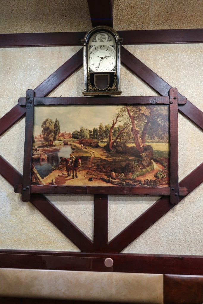 壁に掛けられた振り子時計と田園風景の絵画が映える昭和レトロな喫茶店の一角|木枠の装飾がノスタルジックな空間を演出