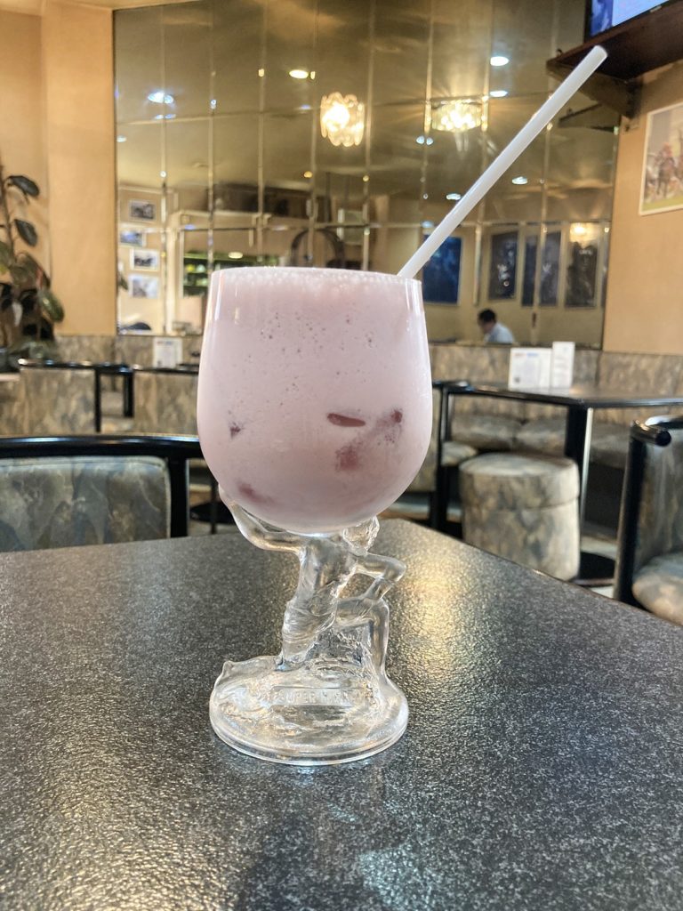 グラスを方に載せて持ち上げる人物型のグラスに入ったピンク色のイチゴヨーグルトドリンク|COFFEE VIPの店内にて