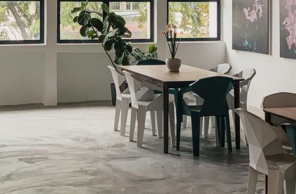 ムラ感のあるモルタル風の床が映えるシンプルモダンなダイニング空間。自然光が差し込む明るい部屋にナチュラルな木製テーブルと個性派チェアが調和