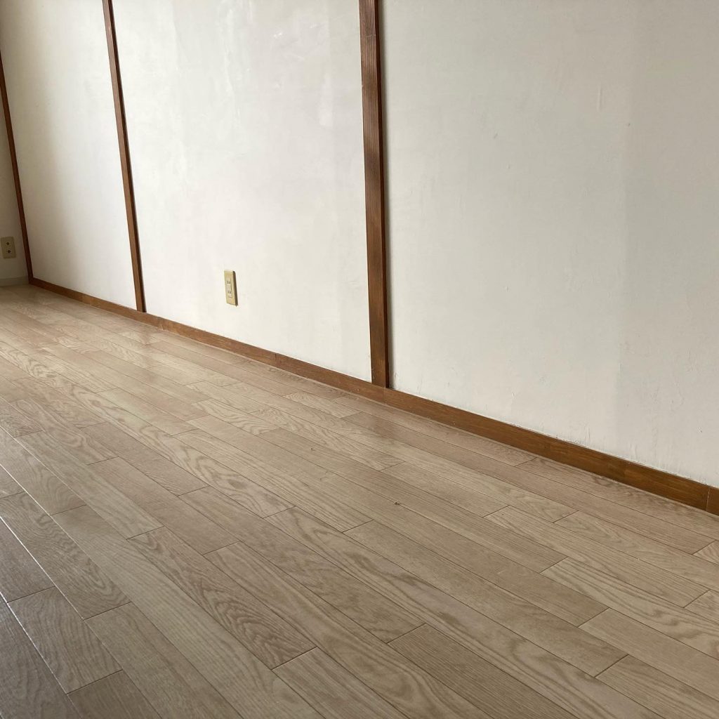 経年で色味が変わった明るいフローリングを壁際から撮影した写真。巾木やコンセント位置も確認できる。