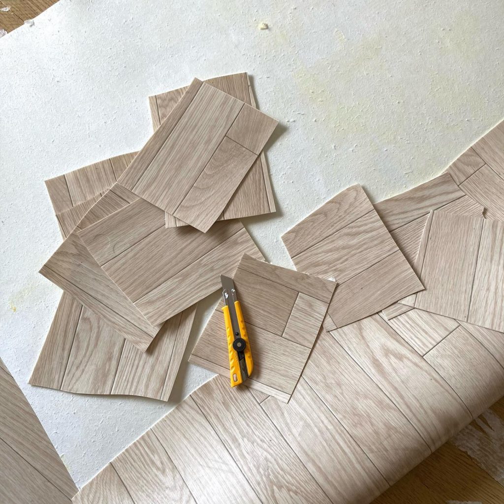 木目柄クッションフロアを細かくカットし、床材の上に並べている作業中の写真。カッターやパーツが散らばっている。