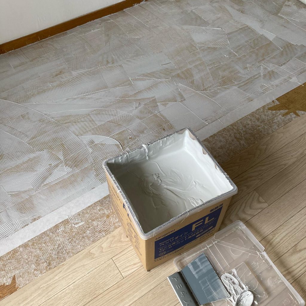 既存床の上に床用接着剤を均一に塗り広げている途中の写真。クッションフロアの一部がめくれている。