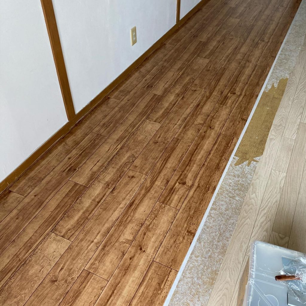 濃い木目柄のクッションフロアを貼り進めている様子。手前が施工済みで、奥に既存床との境目が見えている。