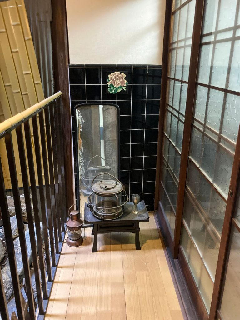 「美香茶楼の室内通路にある黒タイル張りの壁面。花柄の装飾タイルが一枚あしらわれ、下には古いランプや釜、鏡が飾られている。