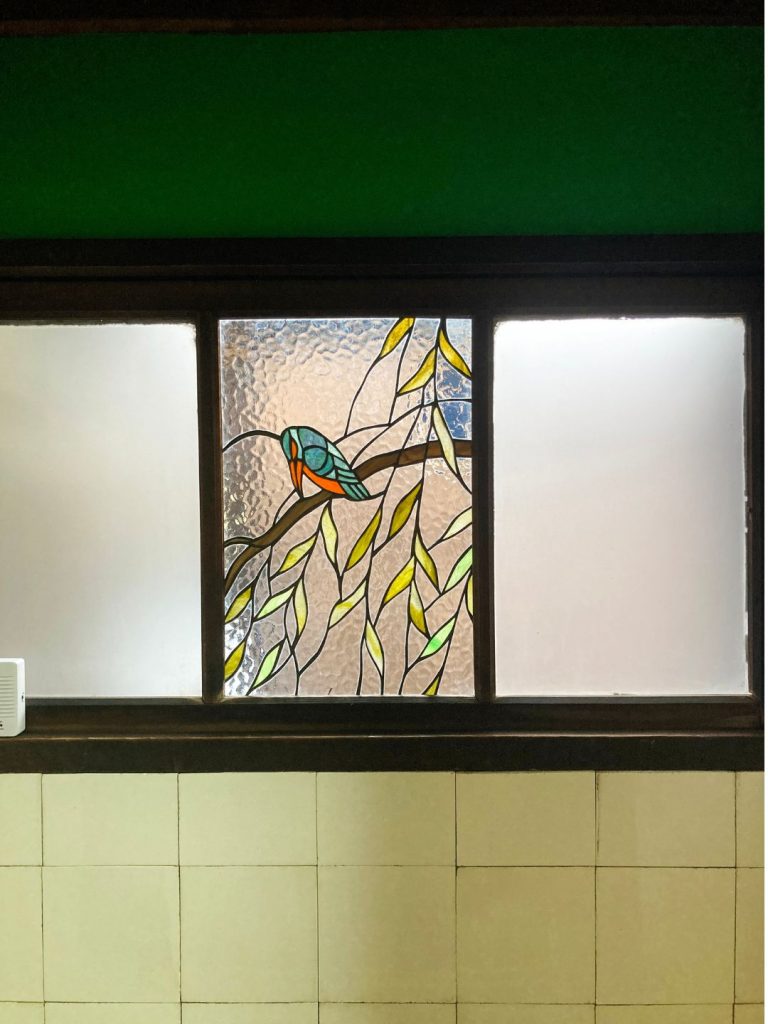 美香茶楼の室内窓に施されたステンドグラス。枝にとまる鳥と揺れる葉を描いた色ガラスが、白いタイル壁の上に鮮やかに浮かび上がる。