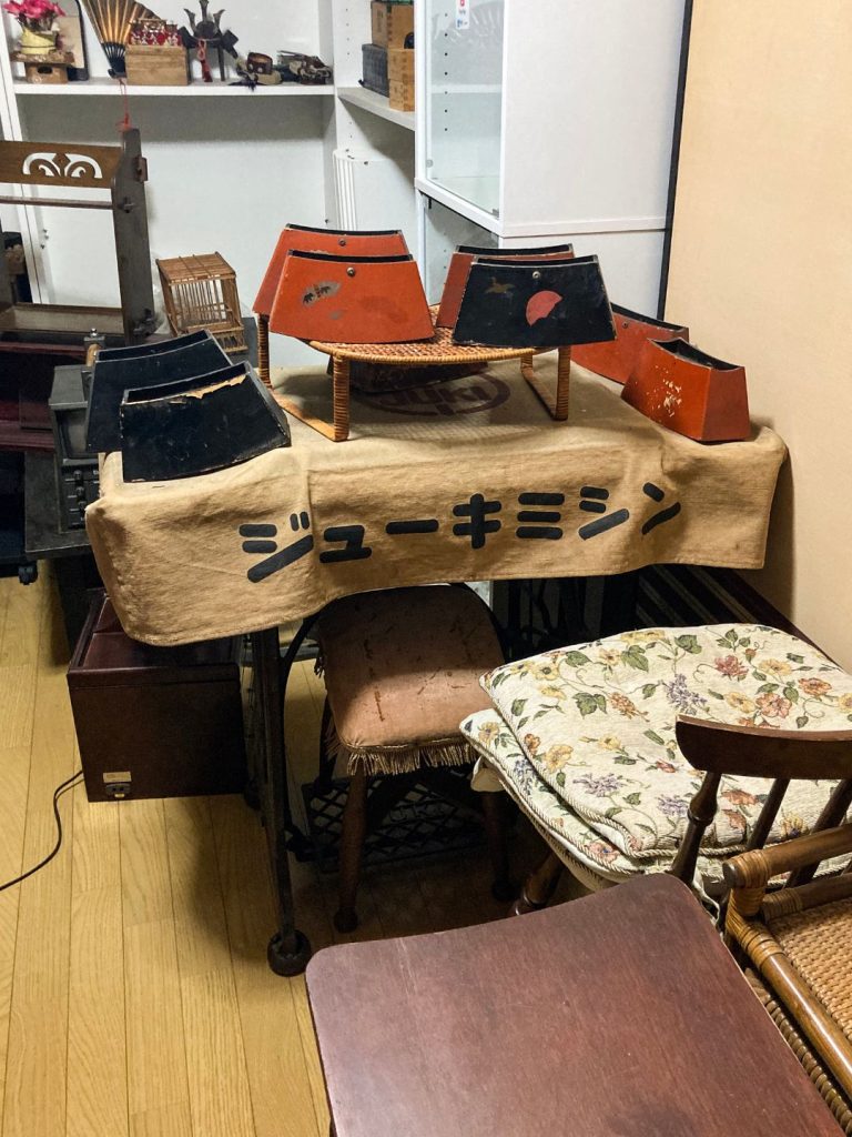 古いジュウキミシン台の上に、朱色と黒色の箱枕の台が並べられている室内の一角。