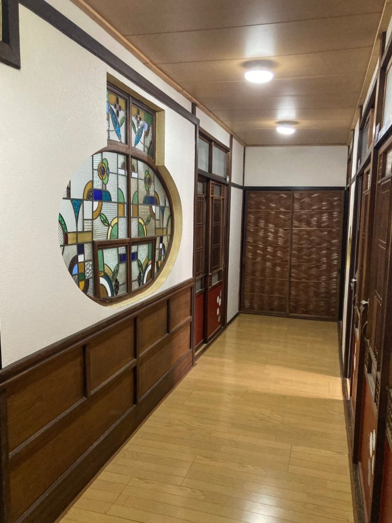 旅館 橋本の香の2階廊下。丸窓に施されたステンドグラスが壁面を彩り、木調の建具と床が調和したレトロな通路。