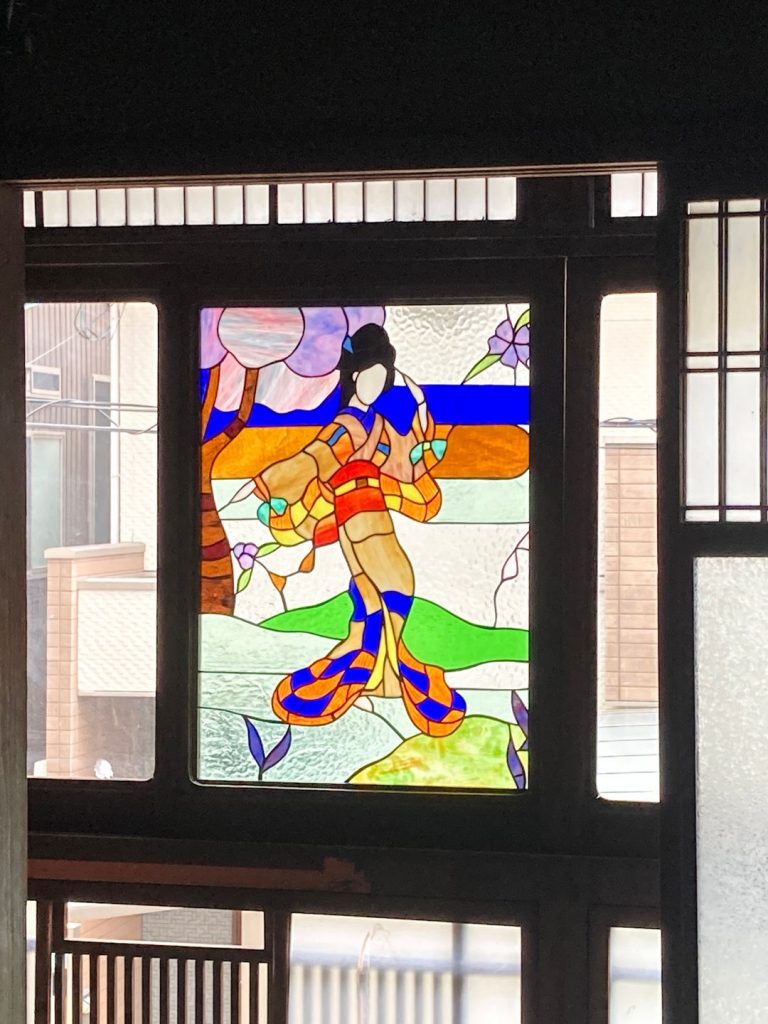 旧加島楼に残る色鮮やかなステンドグラス。着物姿の女性が描かれ、背後には山並みや花木がデザインされた一枚が窓枠にはめ込まれている。