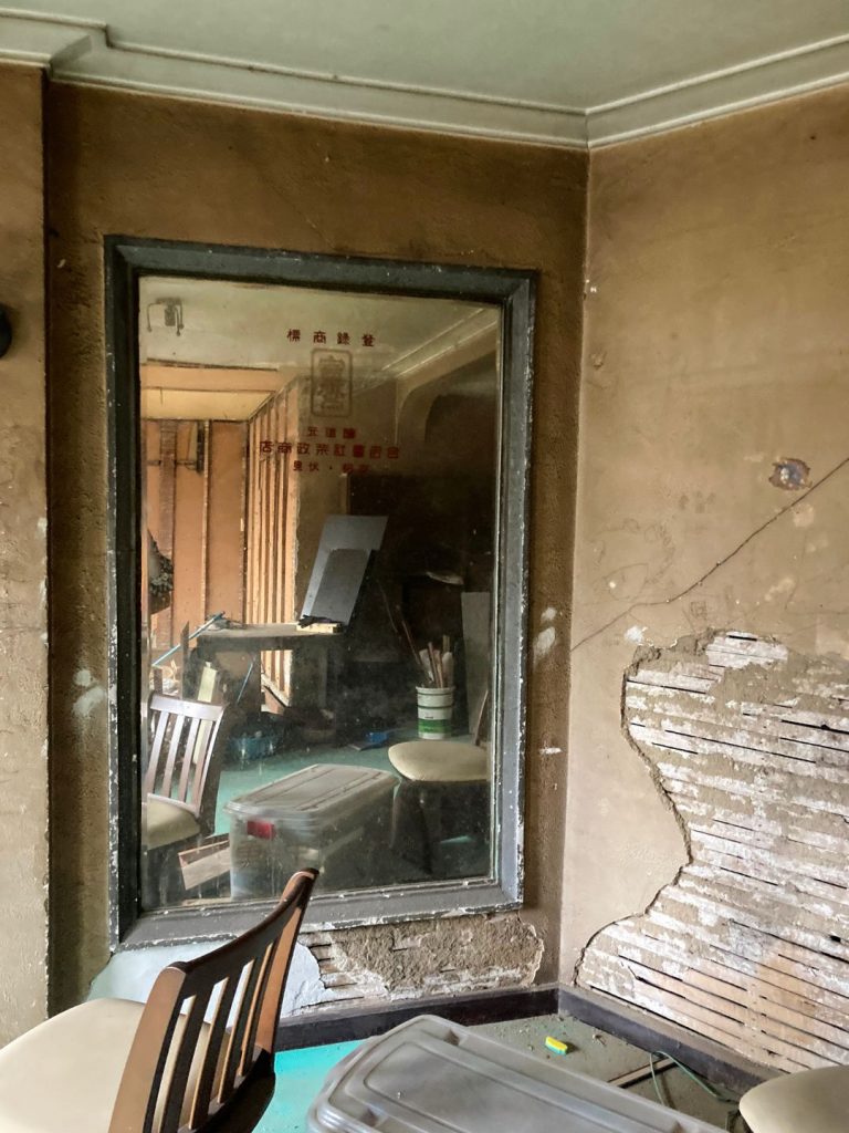 旧加島楼2階の一角に残る大きなガラス窓。剥がれた壁の下地が露出し、室内には椅子や道具類が映り込んでいる様子が写っている。