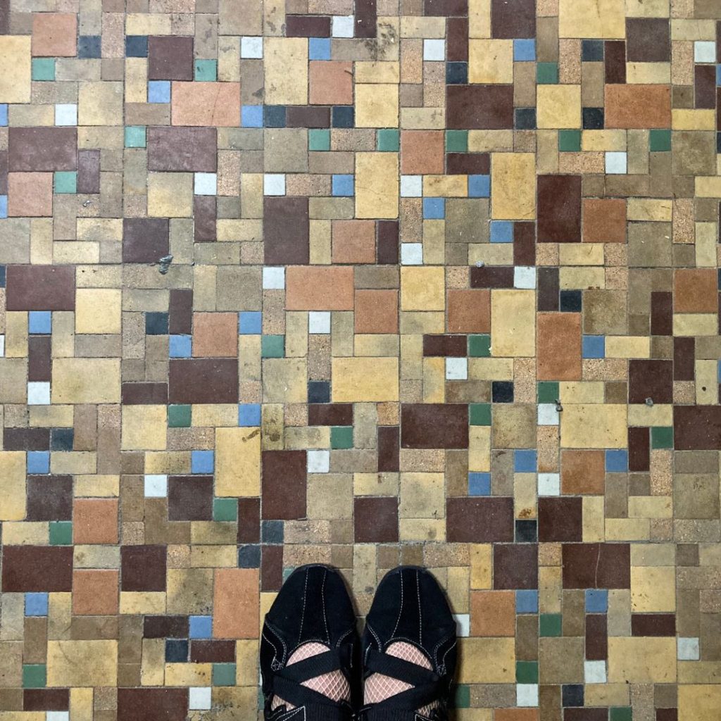 旧加島楼の床。さまざまな色の小さなタイルを不規則に組み合わせたモザイク柄の床と、黒い靴を履いた人物の足先の様子。