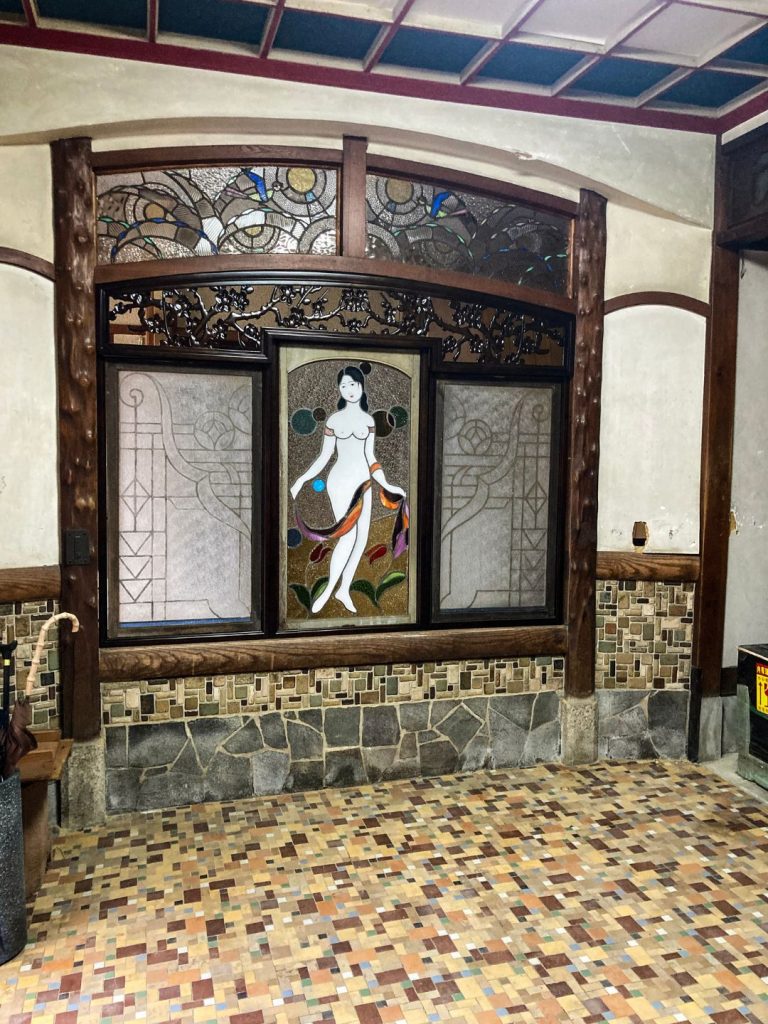 旧加島楼のホールに残るステンドグラス装飾。中央に女性像を描いたステンドグラスと木彫欄間、その周囲をモザイクタイルの床と腰壁が彩るレトロな空間。