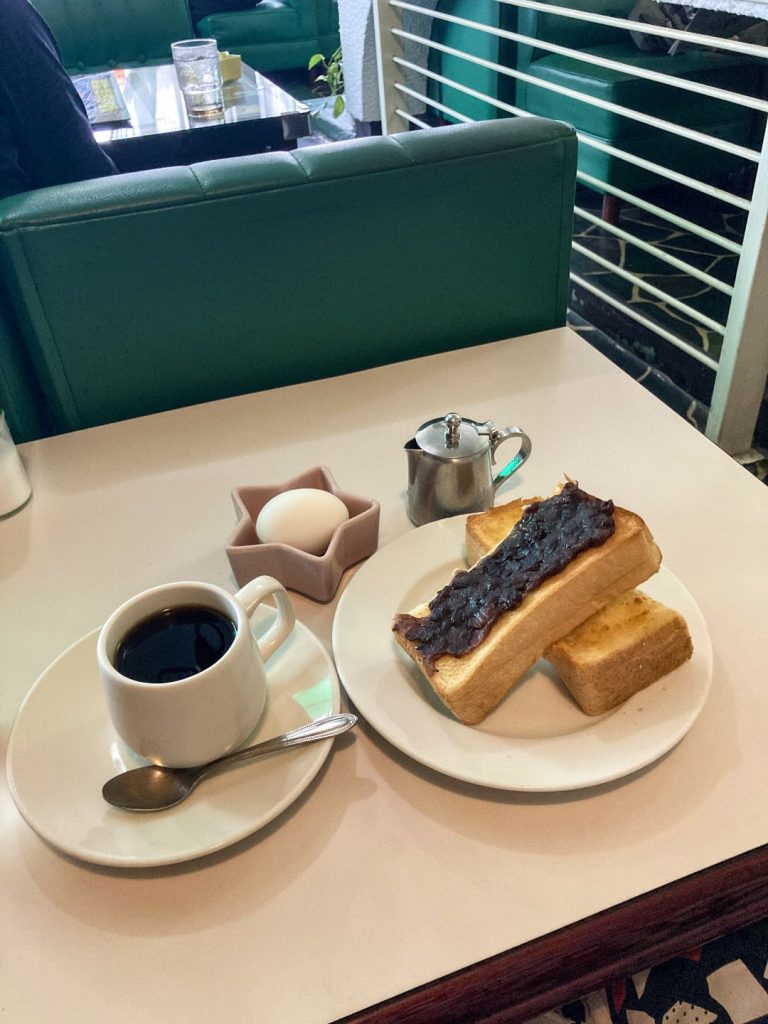 喫茶店のテーブルに並んだモーニングセット。白いカップのブラックコーヒー、あんこをのせた厚切りトースト、ゆで卵、ミルクピッチャーが整然と配置され、奥にはグリーンのソファ席が見える。