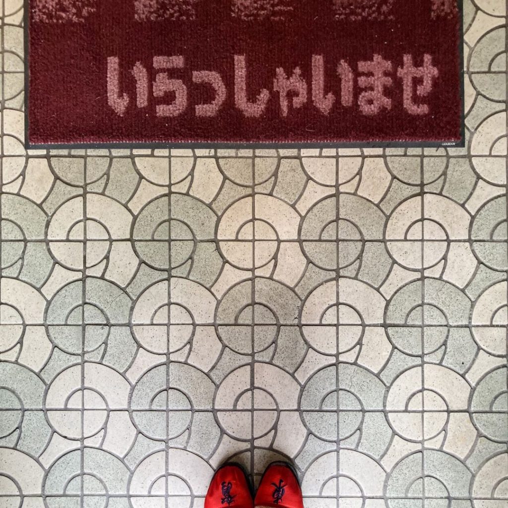 喫茶パスカル青山の店内入口付近の床。淡いグレーとベージュの円形模様が連続するタイル床に、赤い靴と「いらっしゃいませ」と書かれた赤いマットが写っている。
