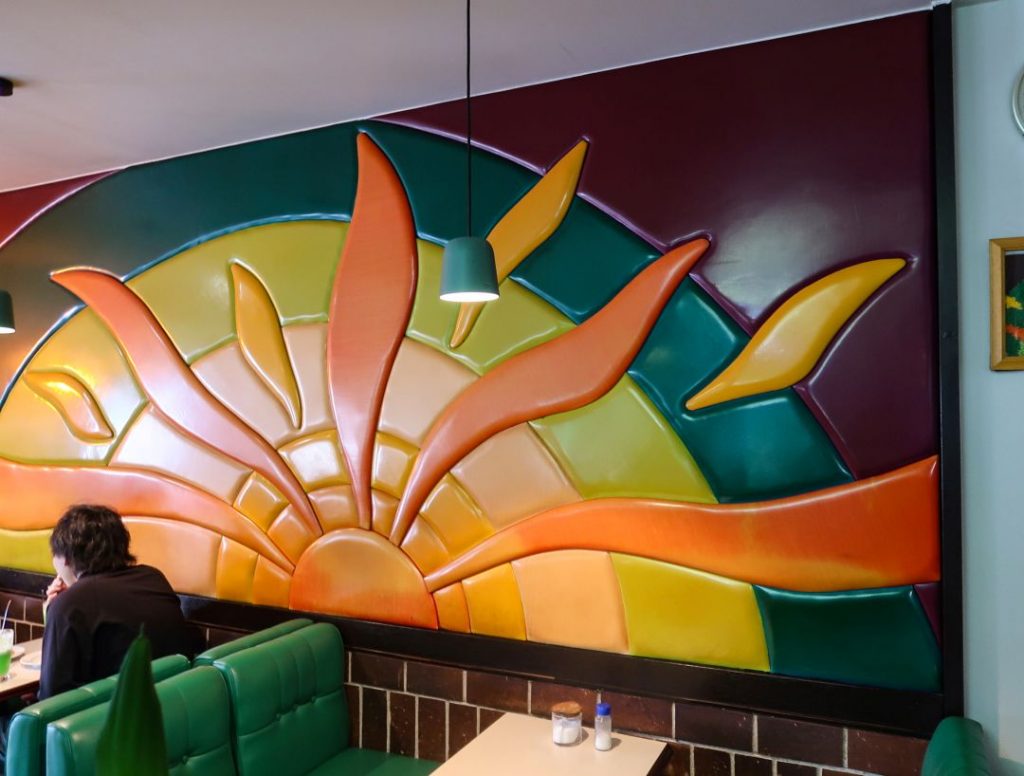 喫茶パスカル青山の店内壁面。オレンジ・イエロー・グリーンを基調にした、朝日や植物を思わせる立体的なレリーフ装飾が広がり、手前にはグリーンのソファ席とテーブルが配置されている。