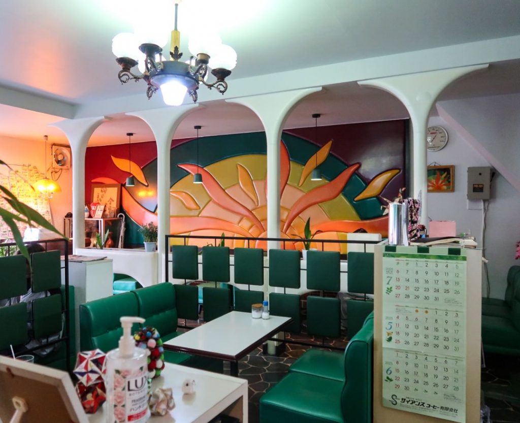 喫茶パスカル青山の店内全景。白いアーチ状の柱越しに、オレンジ・イエロー・グリーンで構成された立体的な壁面レリーフが広がり、手前にはグリーンのソファ席と白いテーブル、天井にはクラシックなシャンデリアが配置されている。