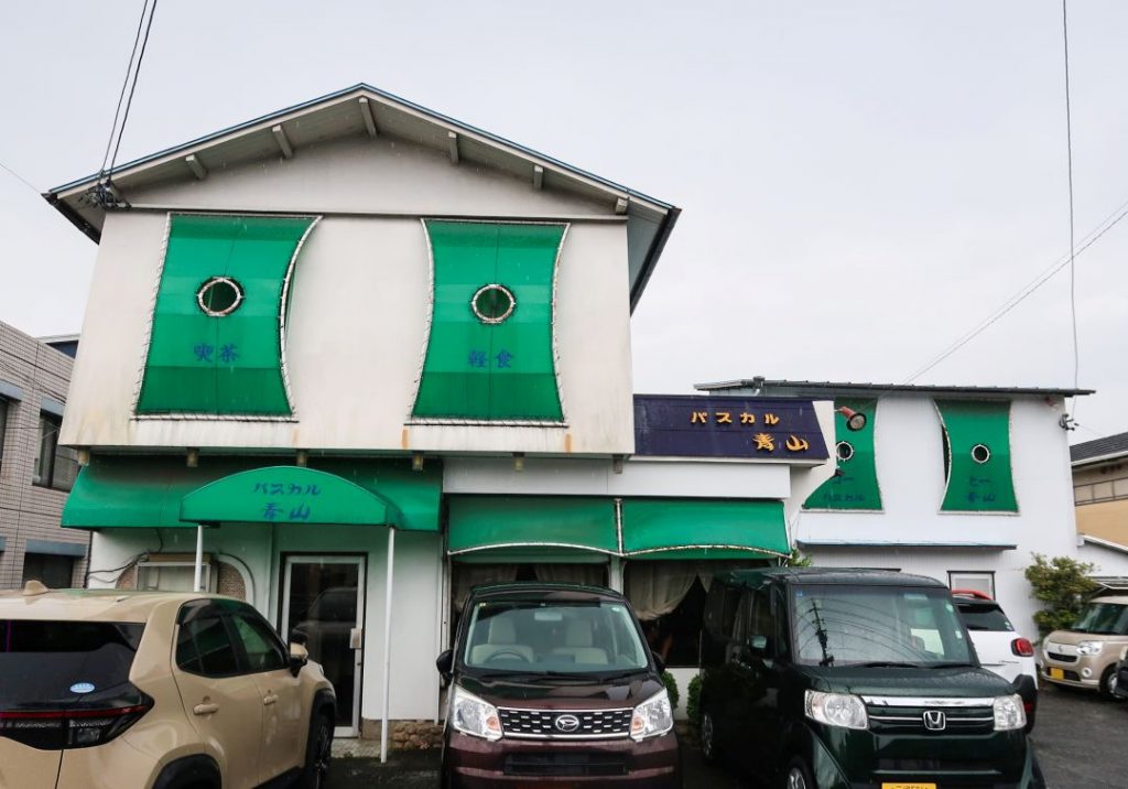 喫茶パスカル青山の外観。白い建物にグリーンの装飾パネルとテントが配され、「パスカル青山」の看板が掲げられている。建物前には駐車スペースがあり、レトロで印象的な佇まいが伝わる。