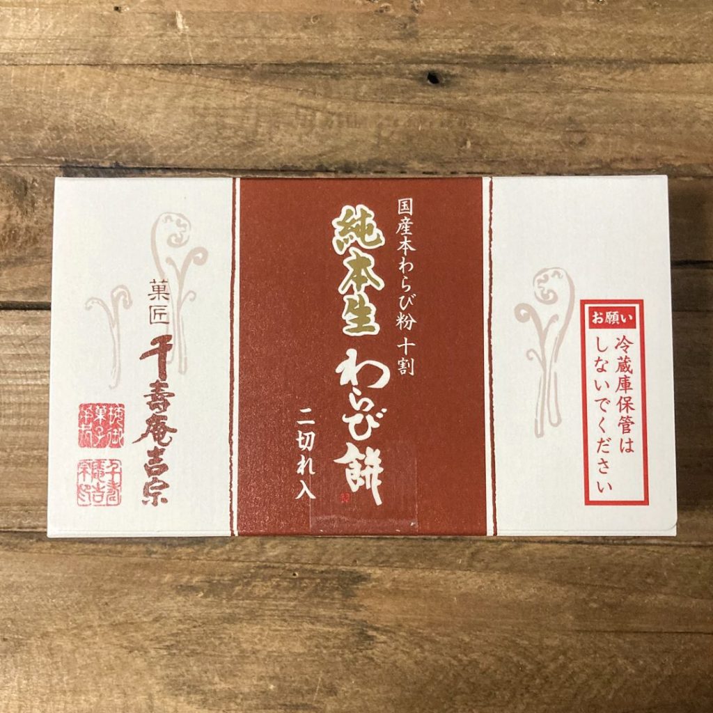 木のテーブルの上に置かれた「純本生わらび餅(二切れ入り)」の箱。赤茶色と白の落ち着いたパッケージに、菓匠「千壽庵吉宗」の文字が入っている。