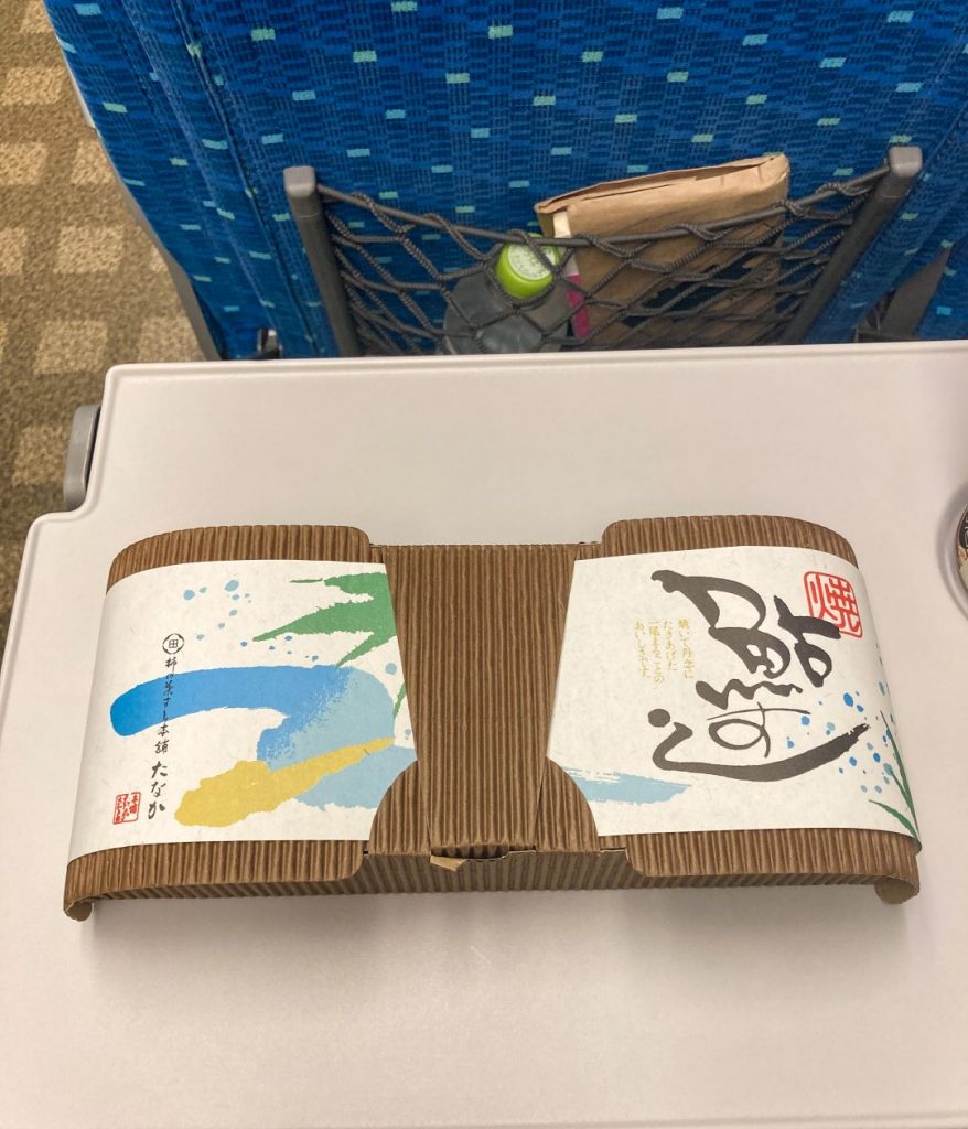 新幹線の座席テーブルに置かれた駅弁。波や筆文字のデザインが施された紙帯で包まれた弁当箱が、車内の青いシートを背景に写っている様子。
