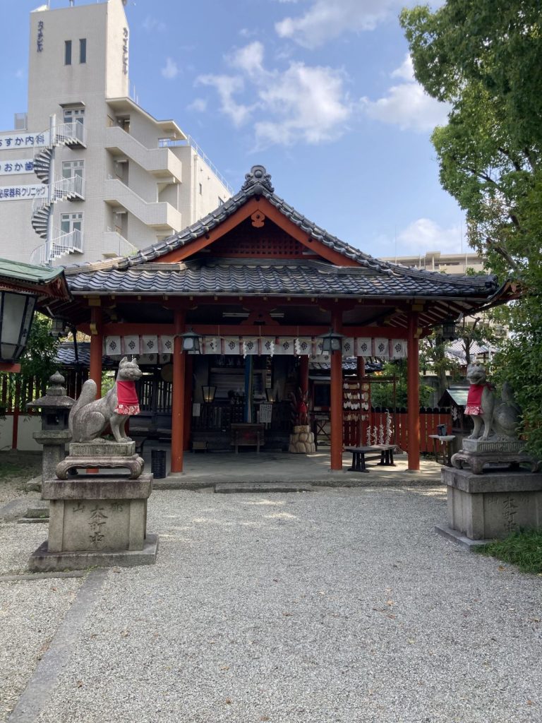 奈良県大和郡山市にある源九郎稲荷神社の境内。朱色の社殿と狛狐が並び、参道正面から社殿を望む風景。