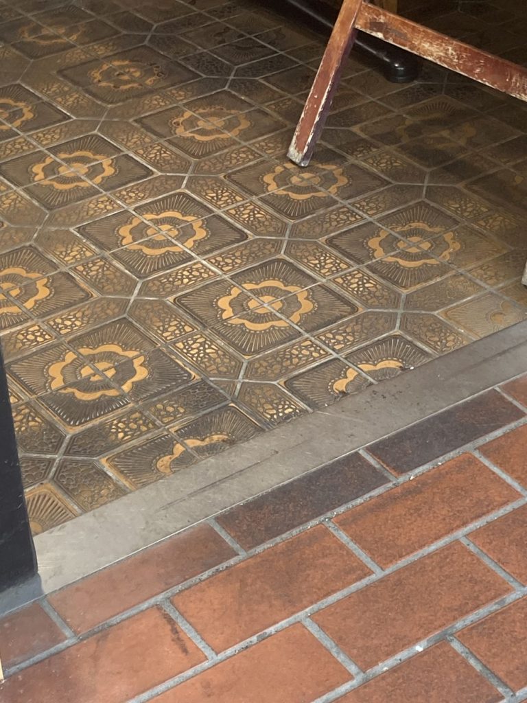 大和郡山商店街の店舗入口付近の床。茶系を基調にしたレトロな幾何学模様タイルが敷かれ、店内の椅子の脚が一部写り込んでいる様子。