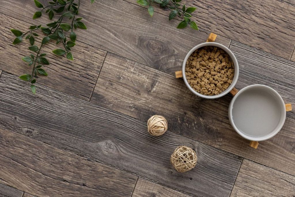 ラグリエのペットマット『エイジドウッド』ヴィンテージ加工を思わせる濃淡のある木目デザインの高機能クッションフロア。古材風の床にエサ入れやインテリア小物が置かれたコーディネート例。