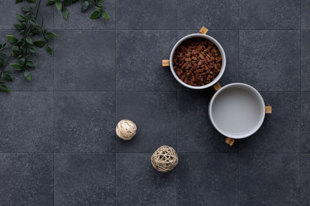 ラグリエのペットマット『グレイブロック』ダークグレーの石目調タイルデザインの高機能クッションフロア。重厚感のある床にエサ入れやインテリア小物が置かれたコーディネート例。