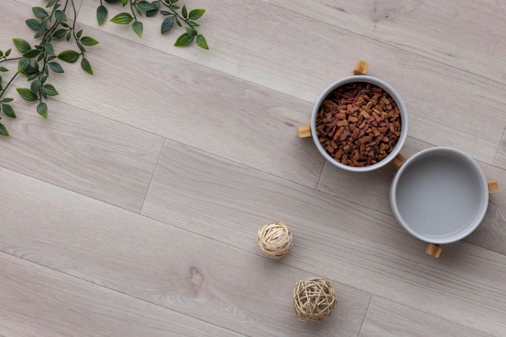ラグリエのペットマット『モイストオーク』淡いグレージュ系の木目デザインの高機能クッションフロア。やさしい色合いの床にエサ入れやインテリア小物が置かれたコーディネート例。