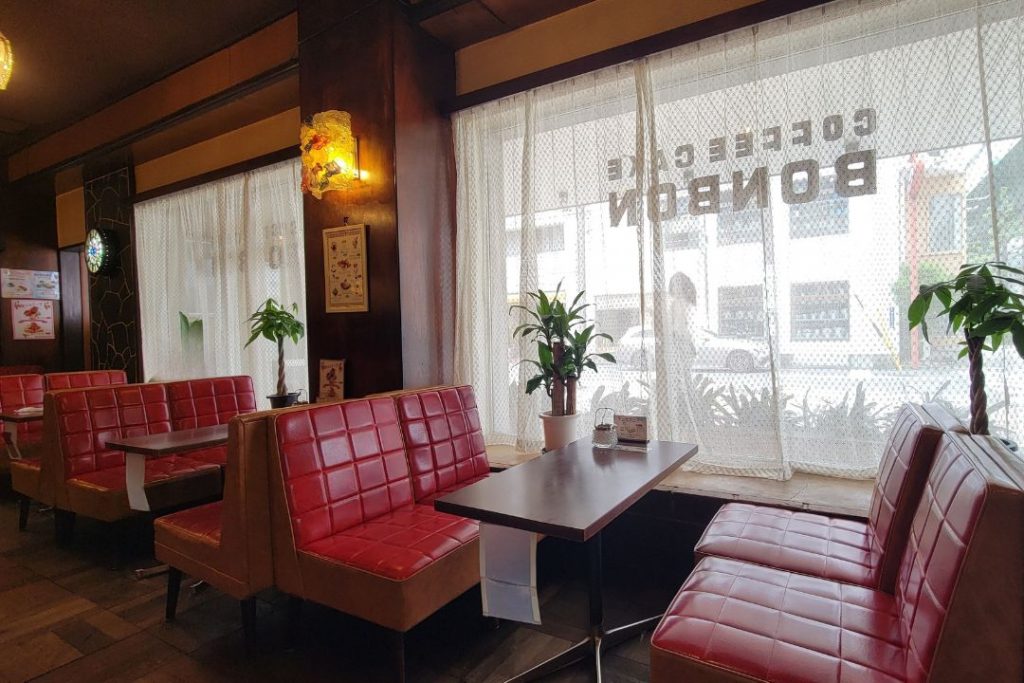 洋菓子・喫茶ボンボンの窓際席。赤いソファとレースカーテン、木目の内装が並ぶ、明るい店内の一角。