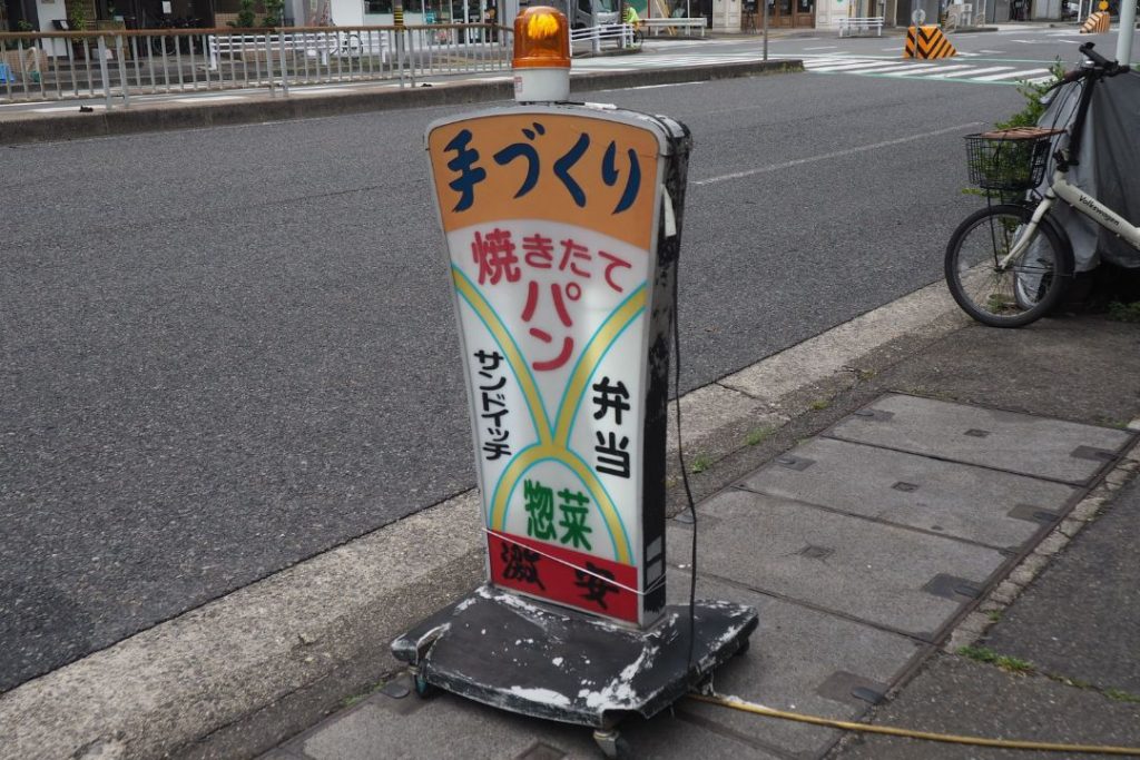 名古屋市東区の街歩きで見つけた手づくりパンの看板。道路沿いに置かれたレトロな立て看板に、焼きたてパンや弁当の文字が描かれている。
